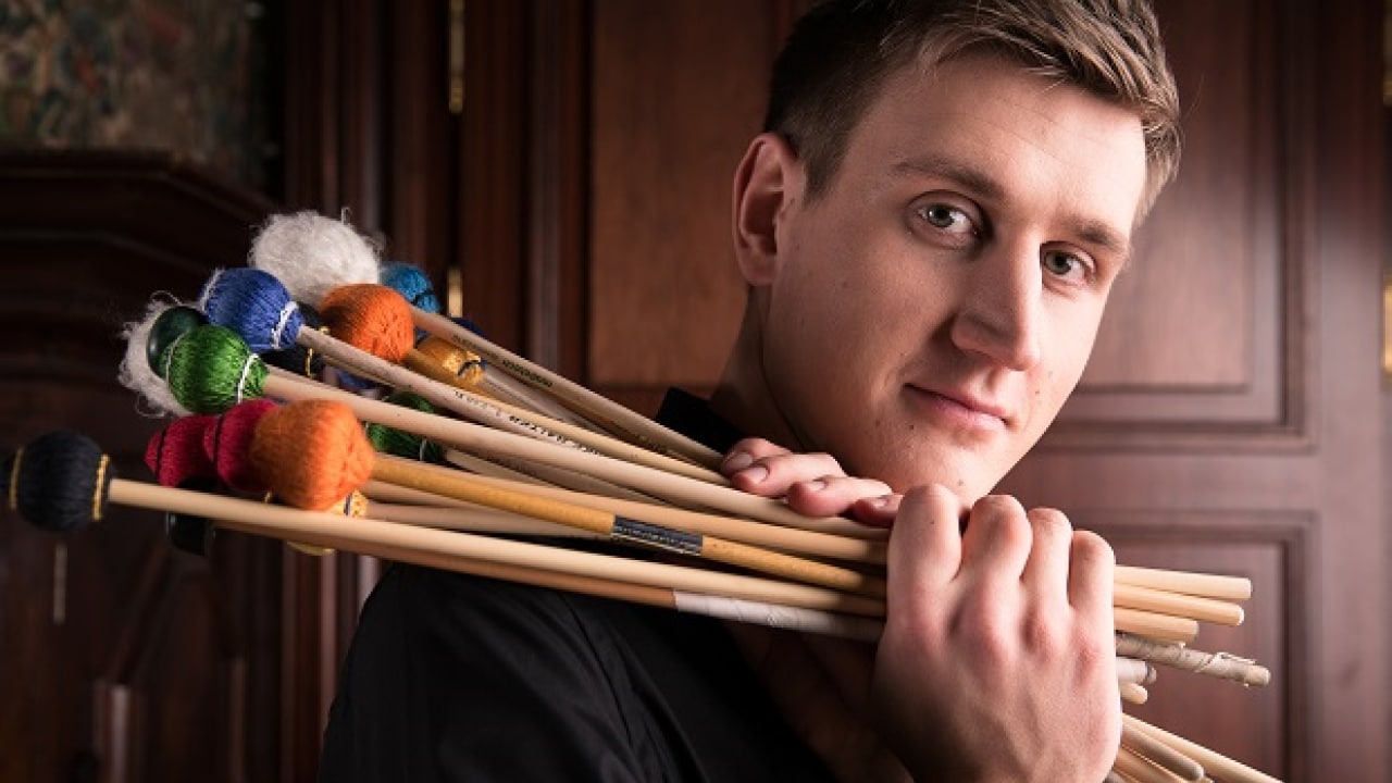 Eine Person mit einer Sammlung von Schlägeln steht vor einem Holzpaneel-Hintergrund.