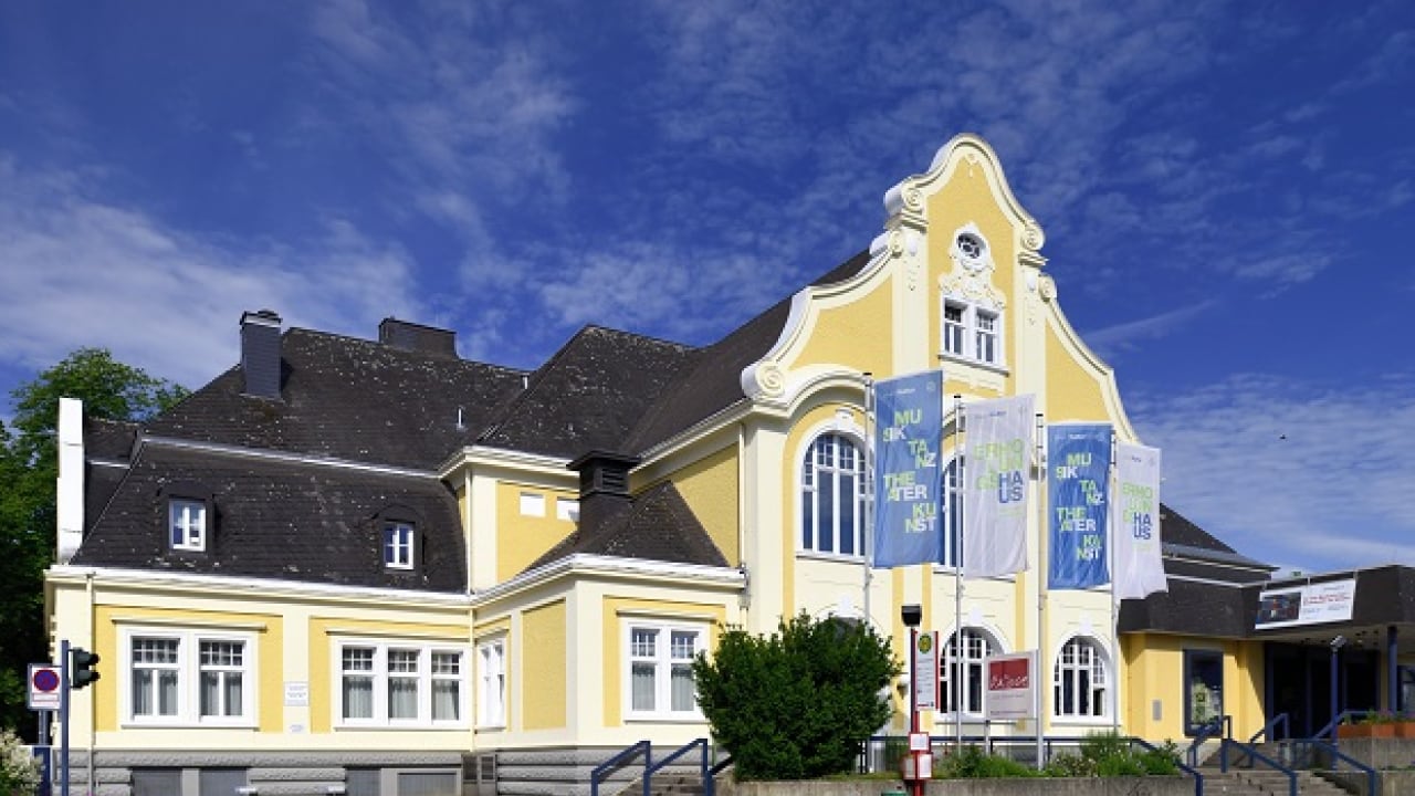 Gelb-weißes Gebäude mit Bogenfenstern und dekorativen Giebeln unter einem blauen Himmel, geschmückt mit mehreren vertikalen Bannern.