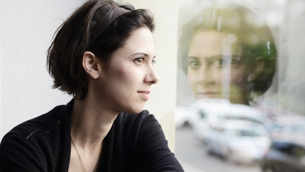 Eine Frau mit kurzen Haaren blickt aus einem Fenster, in dem sich eine sanfte Außenszene mit verschwommenen Autos und Bäumen widerspiegelt.