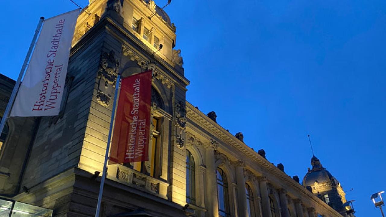 Außenansicht der Historischen Stadthalle Wuppertal, beleuchtet bei Einbruch der Dunkelheit, mit kunstvollen architektonischen Details und zwei Flaggen auf der linken Seite.
