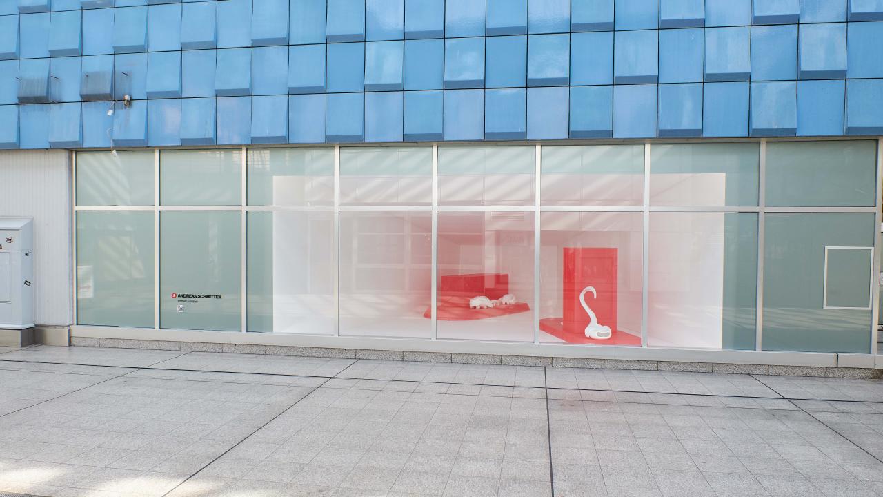 Schaufenster mit großen Glasfenstern und minimalistischer Auslage mit einer weißen Lampe auf einer roten Plattform. Die Außenfassade des Gebäudes ist mit blauen Fliesen verkleidet.