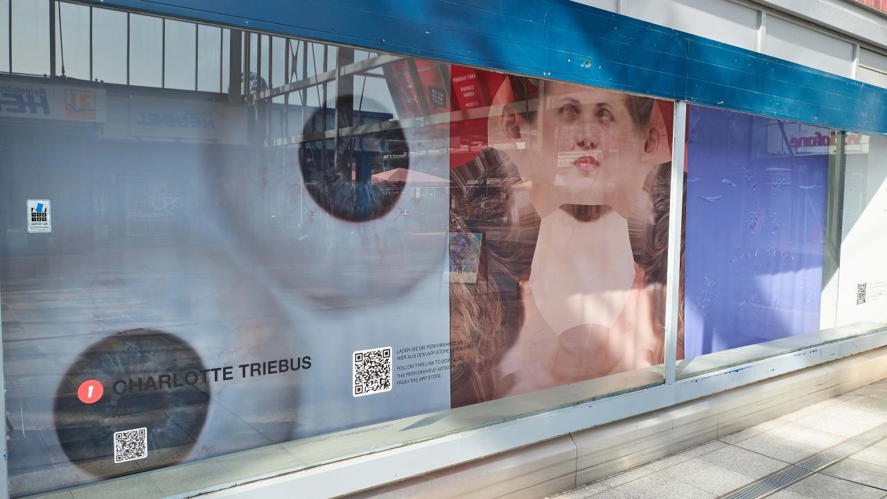 In einem Schaufenster ist ein unscharfes Bild eines Gesichts mit dem Text „Charlotte Triebus“ und QR-Codes zu sehen.