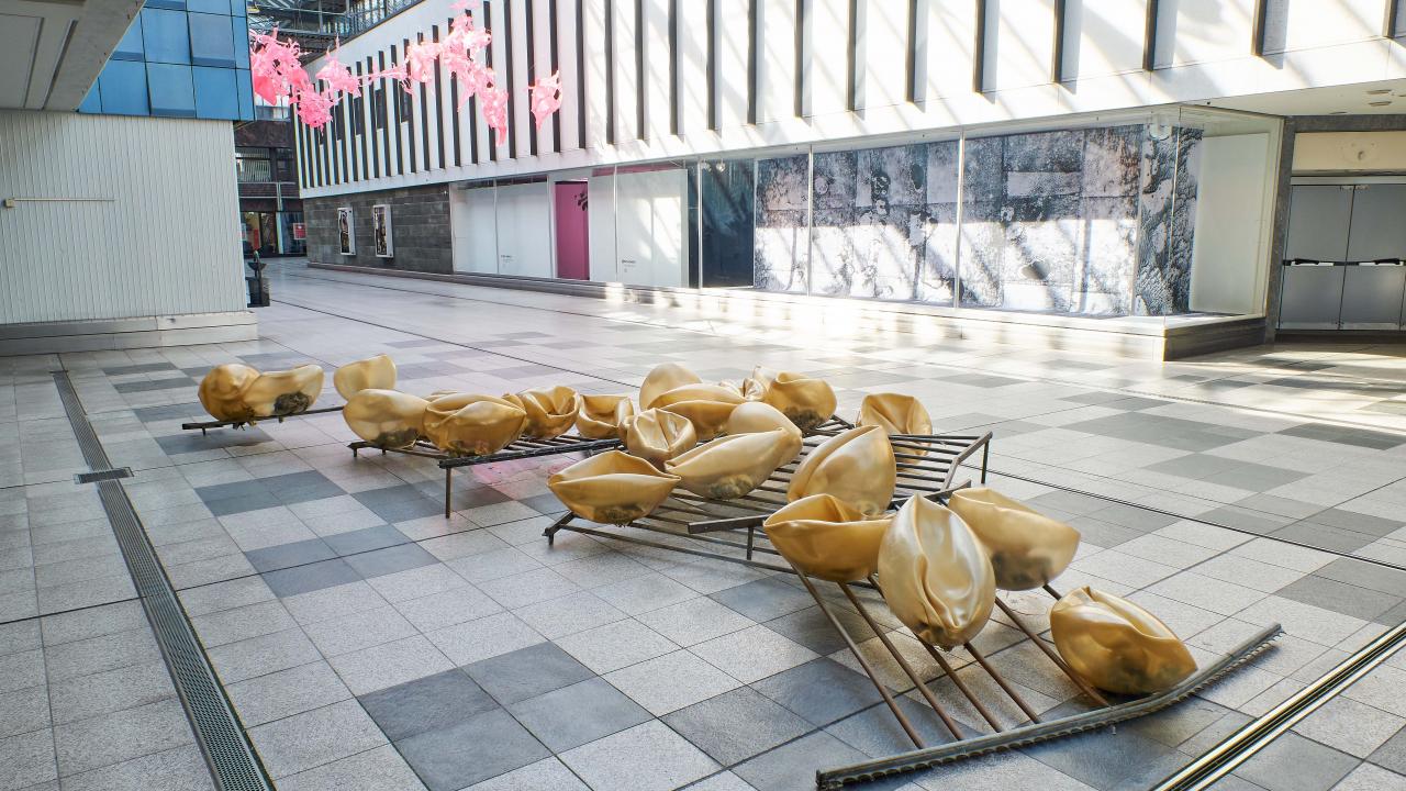 Moderne Kunstinstallation mit großen, goldfarbenen abstrakten Formen auf einem Metallrahmen, ausgestellt in einem geräumigen, gefliesten Innenbereich.