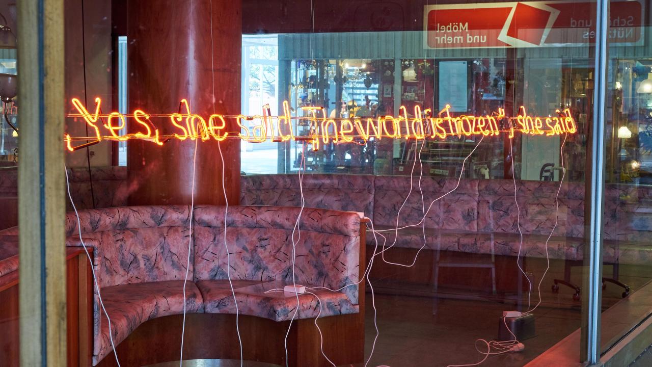 Auf einem Neonschild in einem Fenster steht: „Ja, sie sagte: Die Welt ist gefroren.“ Im Hintergrund ist ein Restaurantstand zu sehen.