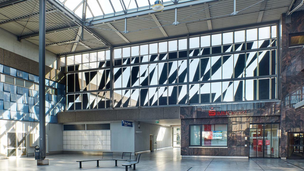 Innenansicht eines modernen Gebäudes mit großen Fenstern, einem Sitzbereich und einem Klinikeingang. Licht strömt durch das gemusterte Glas darüber und schafft eine helle und offene Atmosphäre.