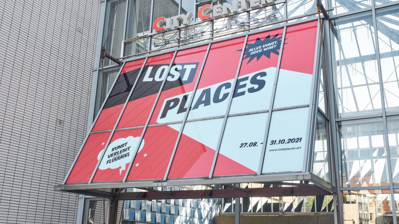 An einem Glasgebäude über einem Eingang mit der Aufschrift „Stadtzentrum“ prangt ein großes Banner mit der Aufschrift „Lost Places“ und den Daten „27.08. – 31.10.2021“.