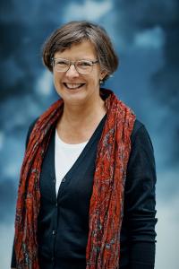 Eine Frau mit kurzen Haaren und Brille trägt eine schwarze Strickjacke, ein weißes Hemd und einen rot gemusterten Schal und steht vor einem verschwommenen blau-weißen Hintergrund. Sie lächelt.