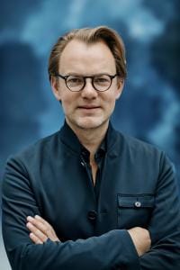 Eine Person mit Brille und dunkler Jacke steht mit verschränkten Armen vor einem blauen Hintergrund.