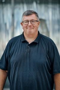 Eine Person mit Brille und dunklem Poloshirt steht lächelnd vor einem unscharfen Hintergrund.