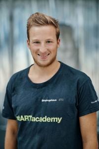 Eine Person mit kurzen Haaren lächelt, trägt ein schwarzes T-Shirt mit dem Aufdruck „stARTacademy“ und steht vor einem unscharfen Hintergrund.
