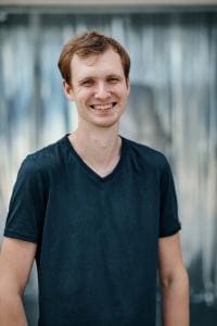 Ein Mann in einem dunklen Hemd steht und lächelt vor einem unscharfen Hintergrund.