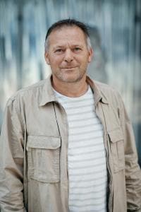 Ein Mann in beiger Jacke und gestreiftem Hemd steht vor einem unscharfen grauen Hintergrund.