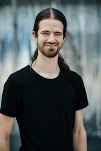 Eine Person mit langen Haaren und Bart und schwarzem T-Shirt lächelt in die Kamera. Der Hintergrund ist unscharf.