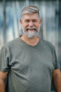 Ein lächelnder Mann mit grauem Haar und Bart sowie einem grauen T-Shirt steht vor einem unscharfen Hintergrund.