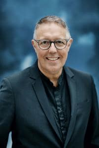 Ein Mann mit Brille und dunklem Anzug lächelt vor einem unscharfen blauen Hintergrund.