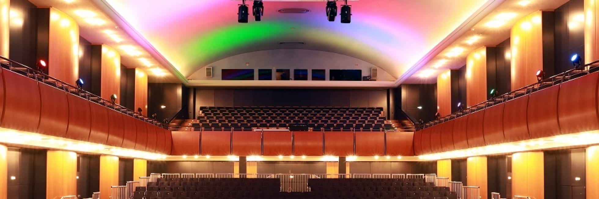 Innenansicht eines leeren Theaters mit beleuchteter Decke und Balkonen, Sitzreihen und einer Bühne. Mehrfarbige Lichter werfen ein Licht von oben.