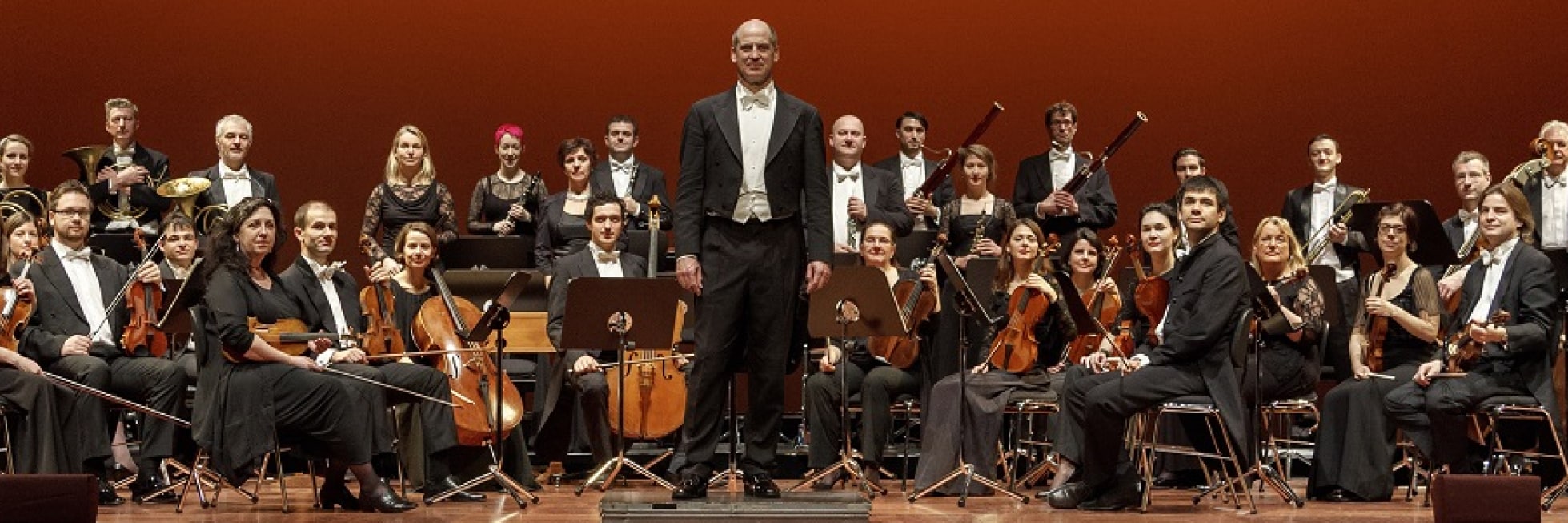 Ein Symphonieorchester auf der Bühne, mit Musikern, die um den Dirigenten herum sitzen und stehen und vor einem dunkelroten Hintergrund verschiedene Instrumente halten.