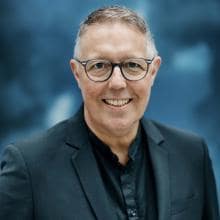 Ein Mann mit Brille und dunklem Anzug lächelt vor einem unscharfen blauen Hintergrund.